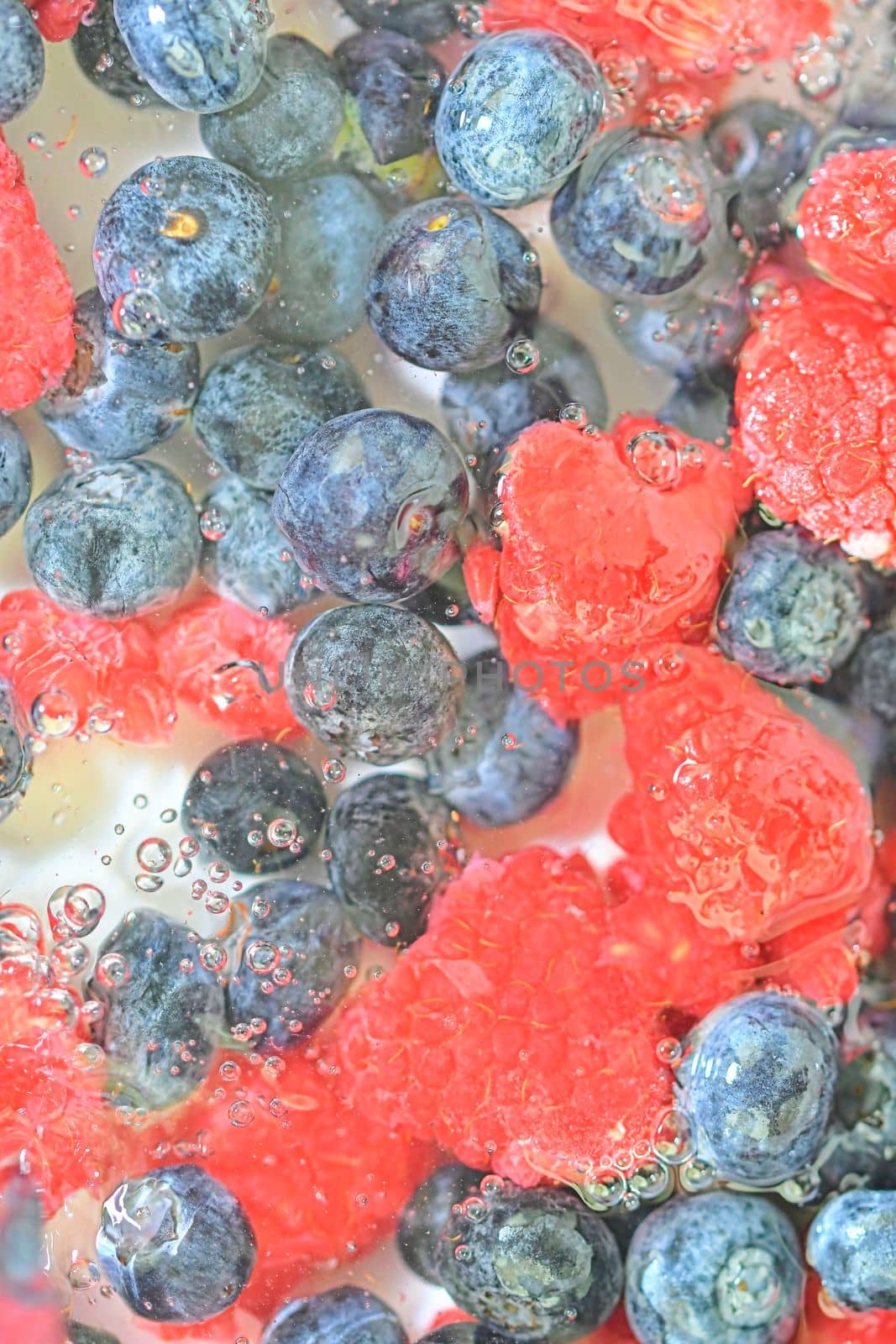 Blueberries and raspberries in liquid with bubbles. Colourful ripe bilberries and raspberries in water. Close-up of fresh berries in water background. Top view, flat design. Vertical macro image.