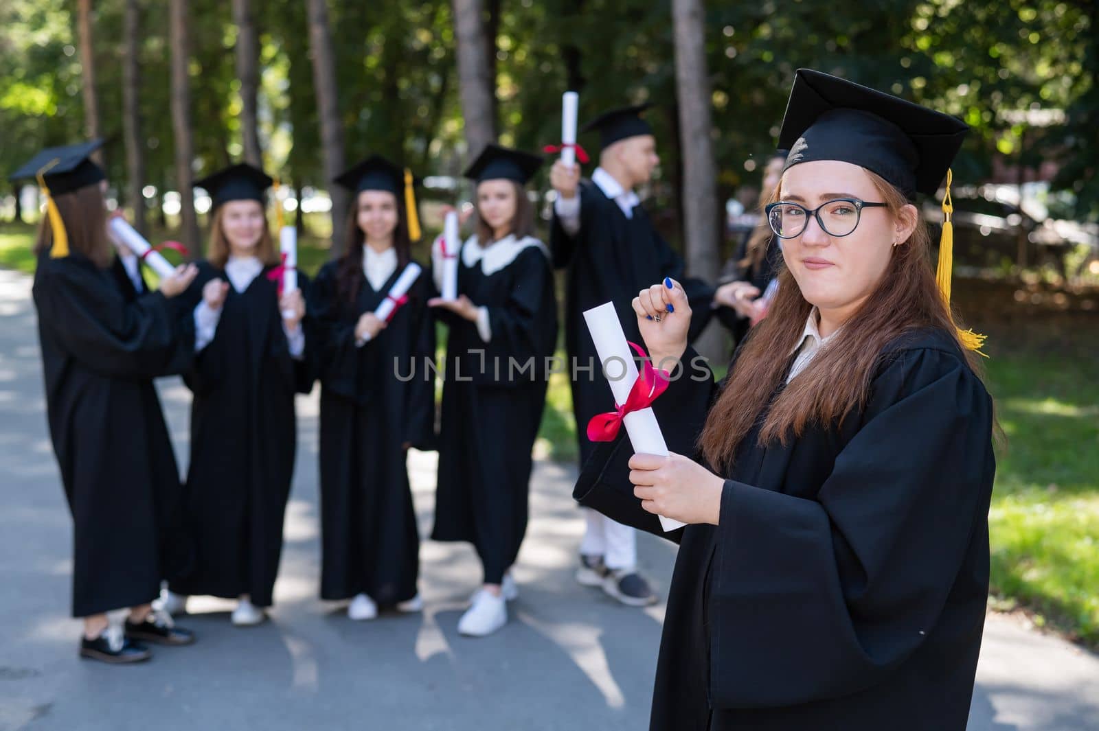 Portrait of a young caucasian woman in glasses and a graduate gown against the background of classmates. A group of graduate students outdoors