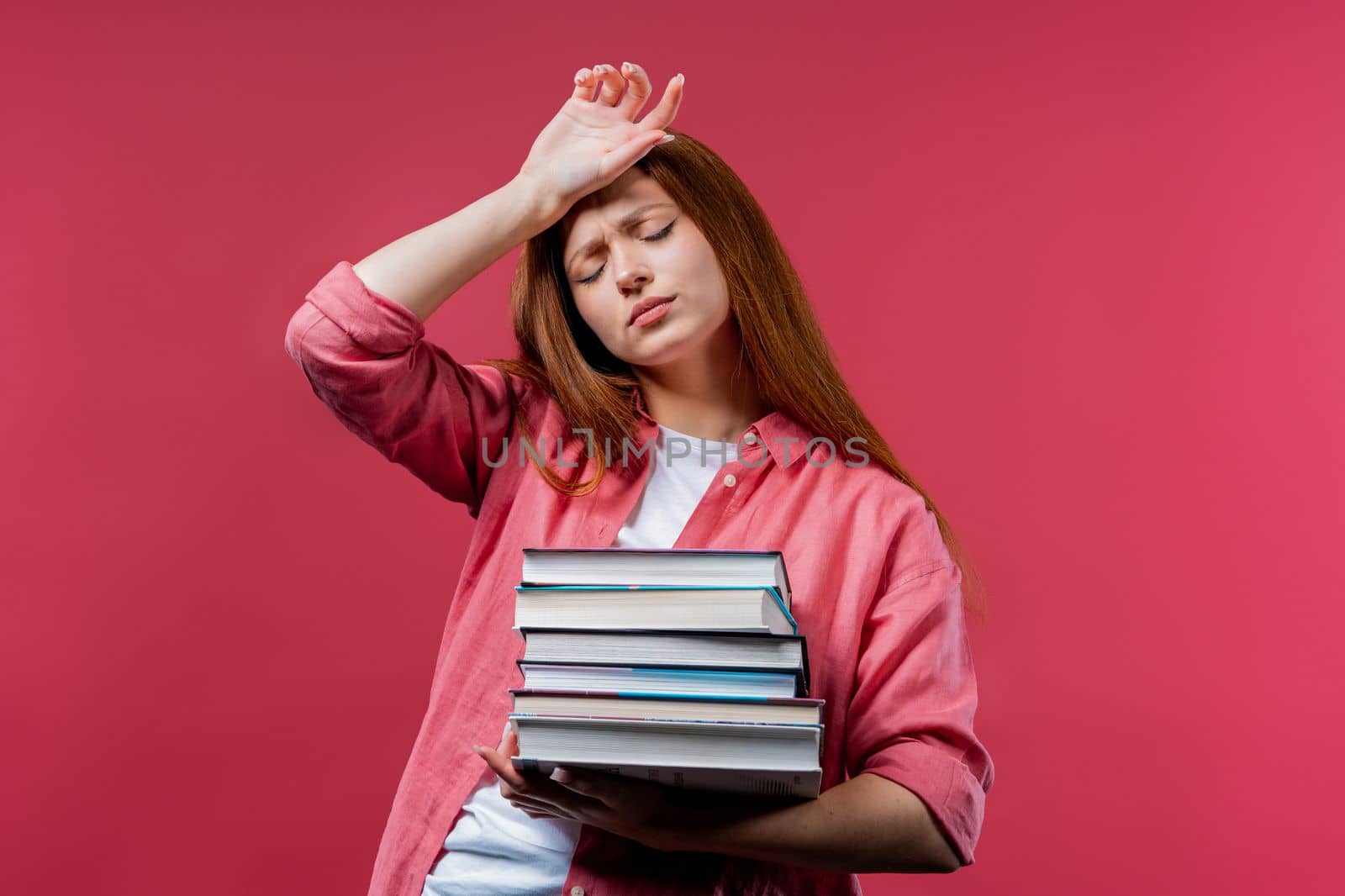 Lazy schoolgirl or student is dissatisfied with amount of books homework on pink background. Girl with ginger hair in displeasure, she is annoyed, discouraged frustrated by studies. High quality photo