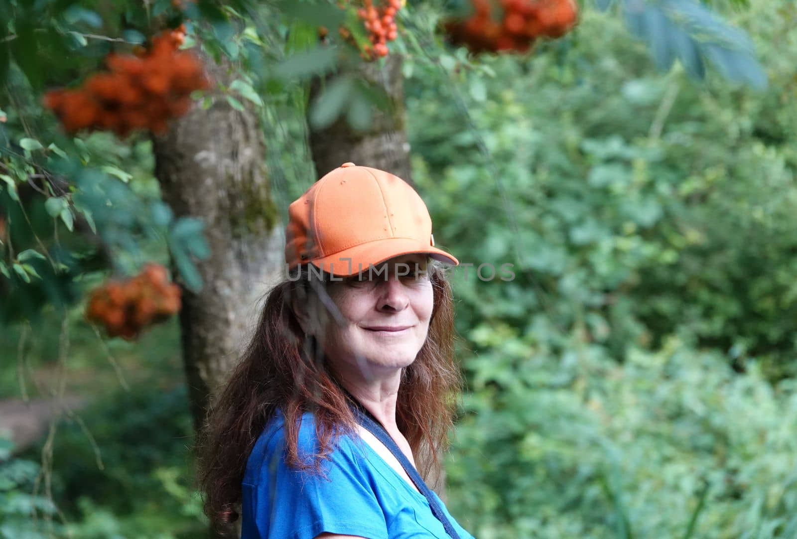 Elderly woman with orange cap beneath blurred orange berry clusters of a rowan tree