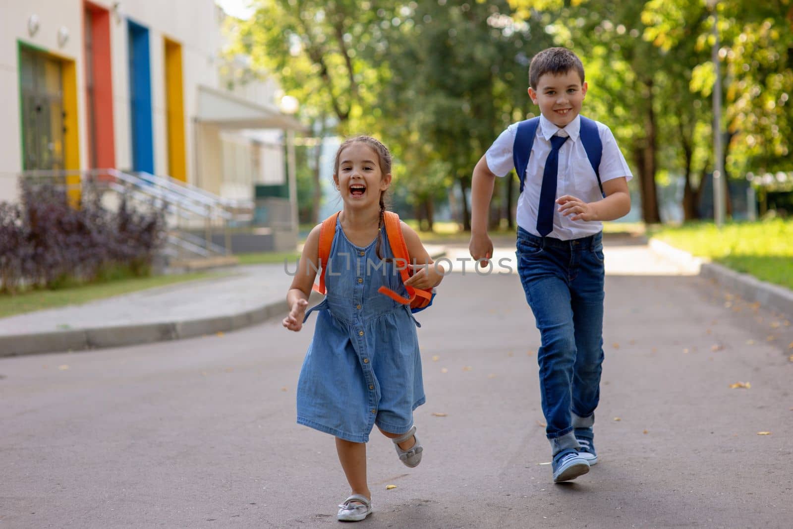Cheerful schoolchildren, a girl and a boy in a white shirt with backpacks run in the morning on a sunny day in summer from school against the background of a school building with colorful windows.