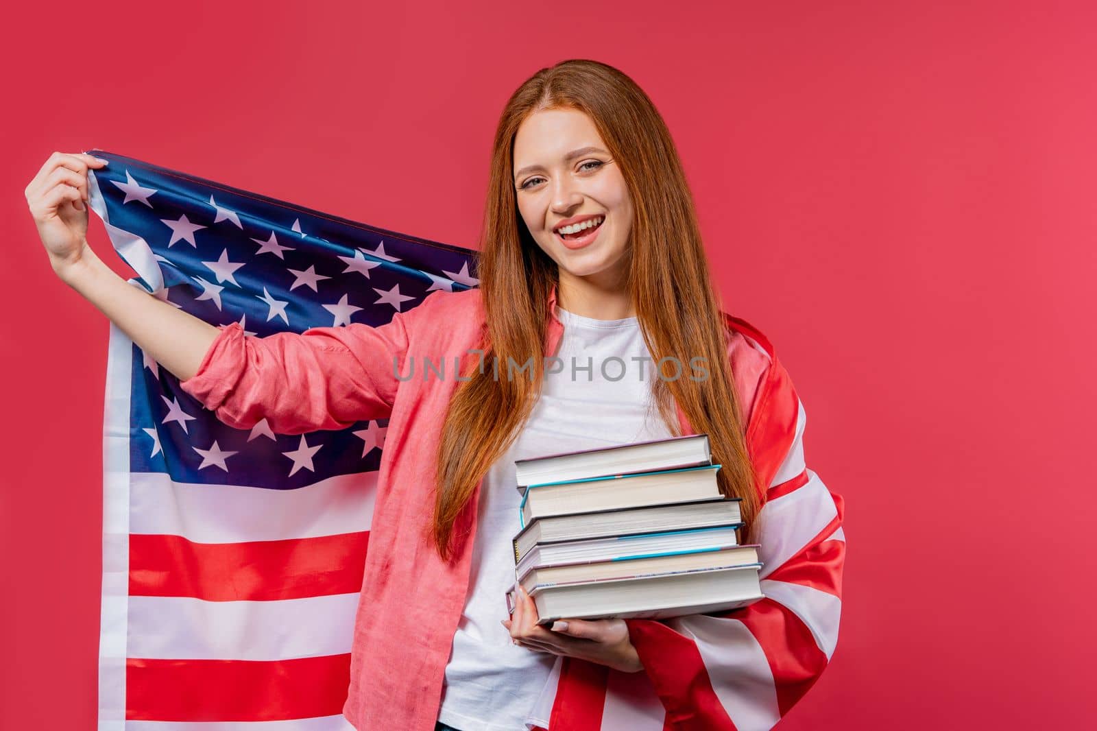 American woman student holds stack of university books from college library on pink background. Happy girl smiles, she is happy to graduate in USA, education abroad concept. High quality photo