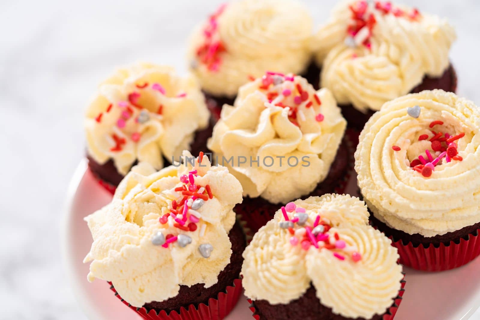 Freshly baked velvet cupcakes with white chocolate ganache frosting decorated with sprinkles.