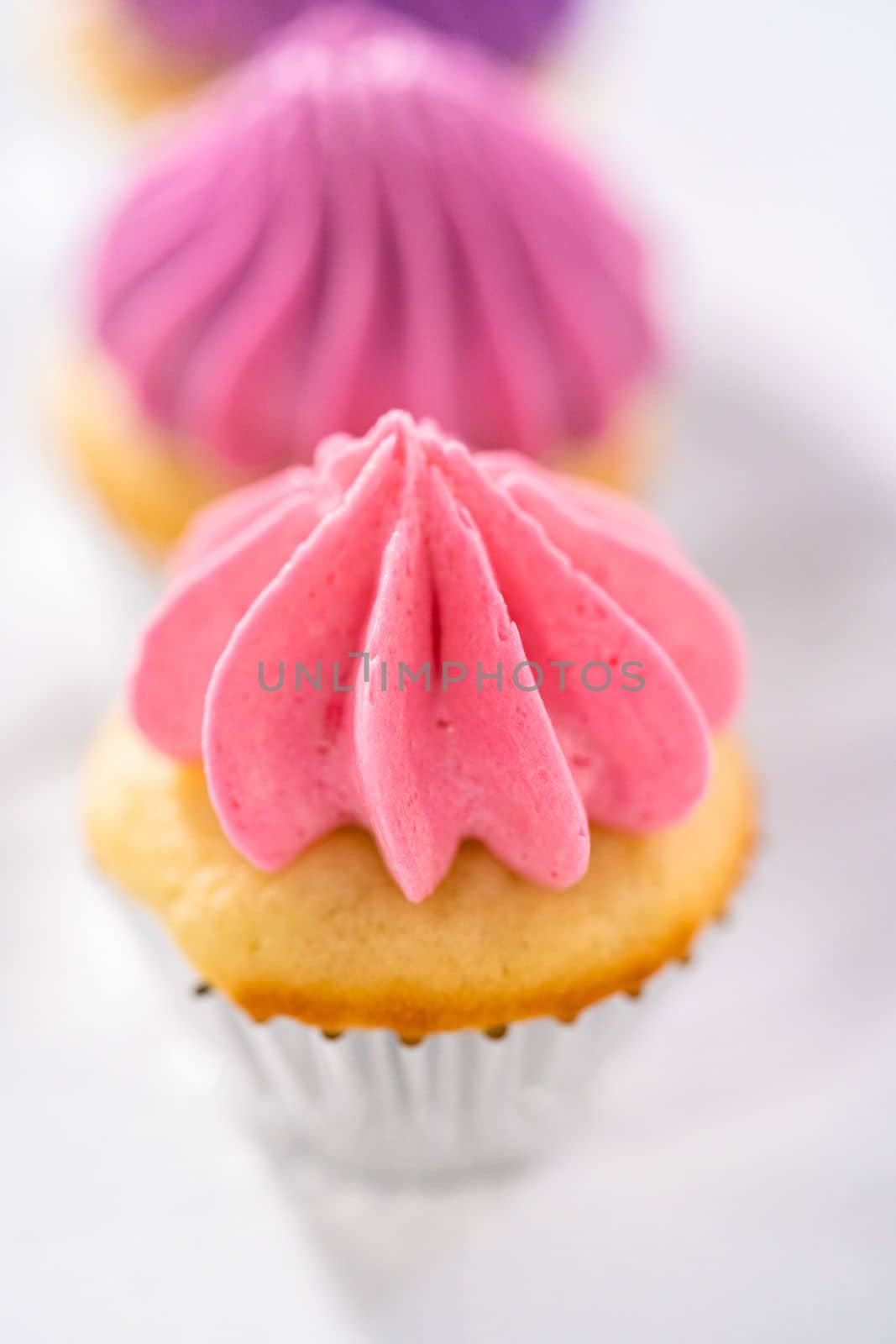 Freshly baked mini vanilla cupcakes with ombre pink buttercream frosting on a marble kitchen ocunter.