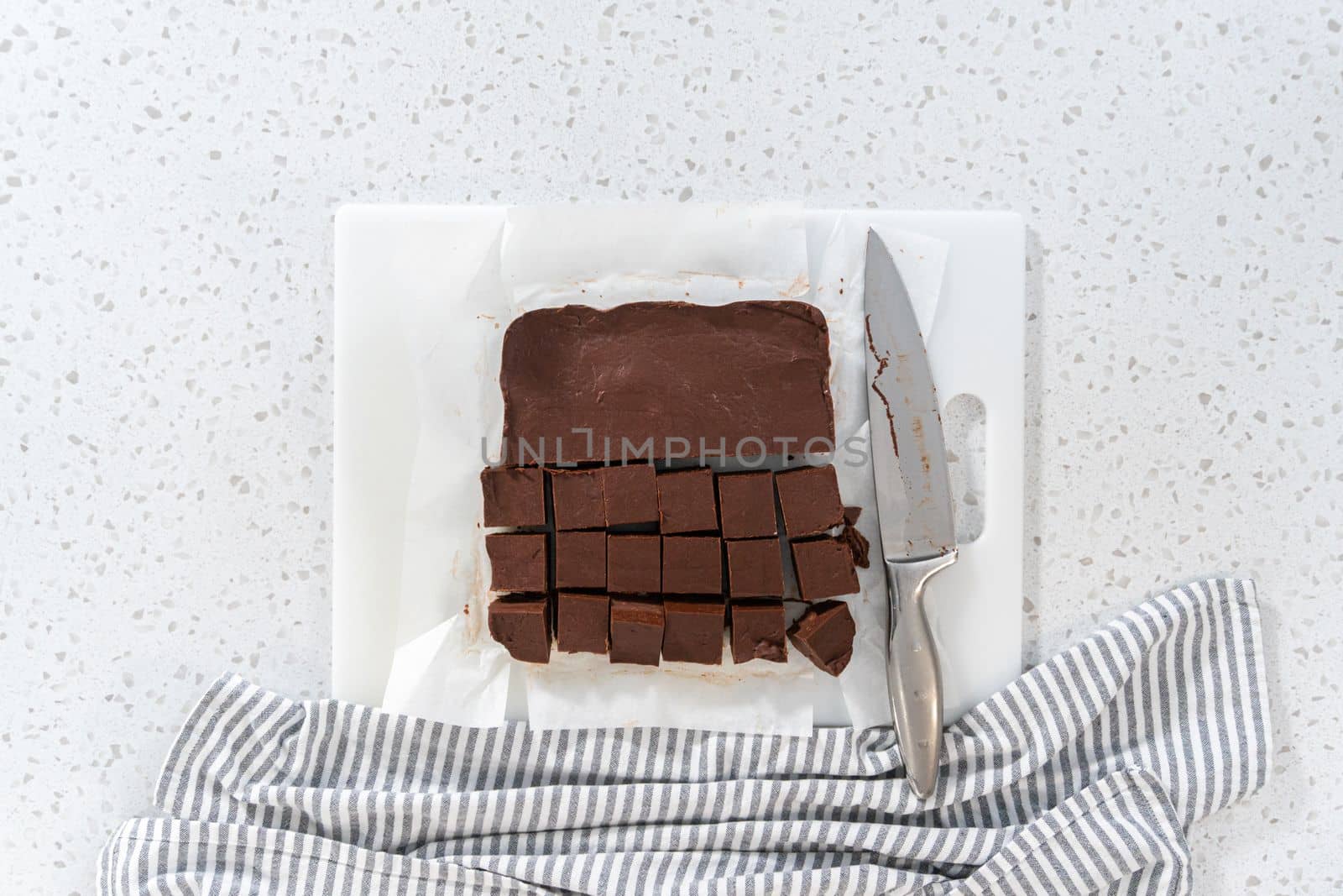 Flat lay. Cutting plain homemade fudge into small pieces on a white cutting board.