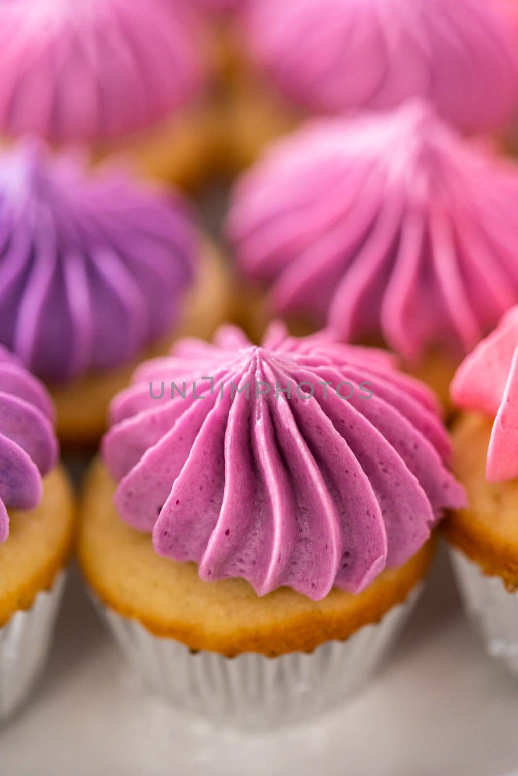 Freshly baked mini vanilla cupcakes with ombre pink buttercream frosting on a white cake stand.