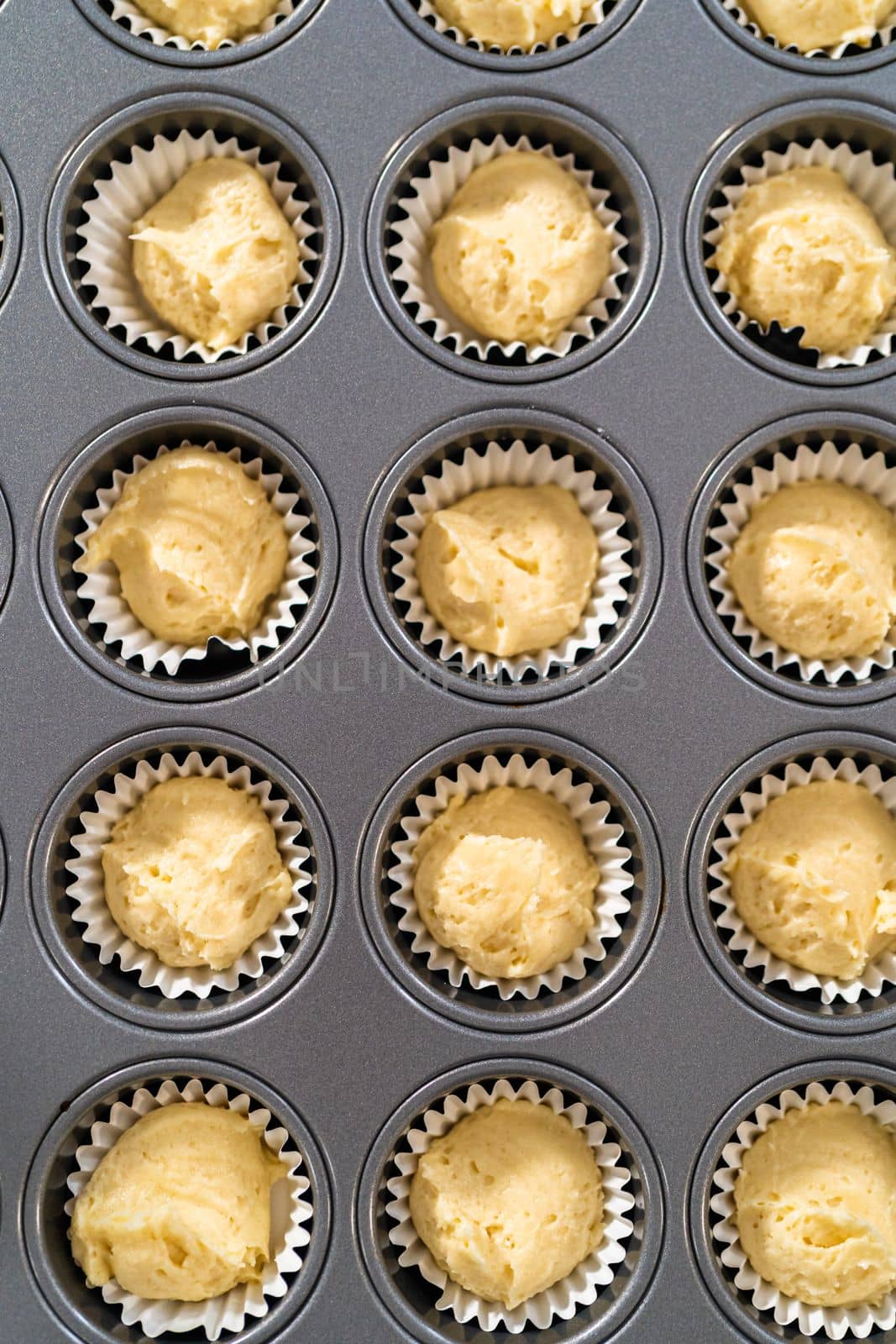 Scooping cupcake batter with dough scoop into a baking cupcake pan with liners to bake mini vanilla cupcakes with ombre pink buttercream frosting.
