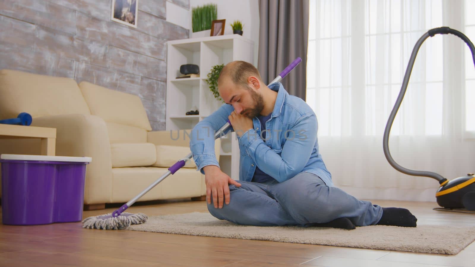 Tired man sitting on cozy carpet after cleaning the room floor with mop and detergent.