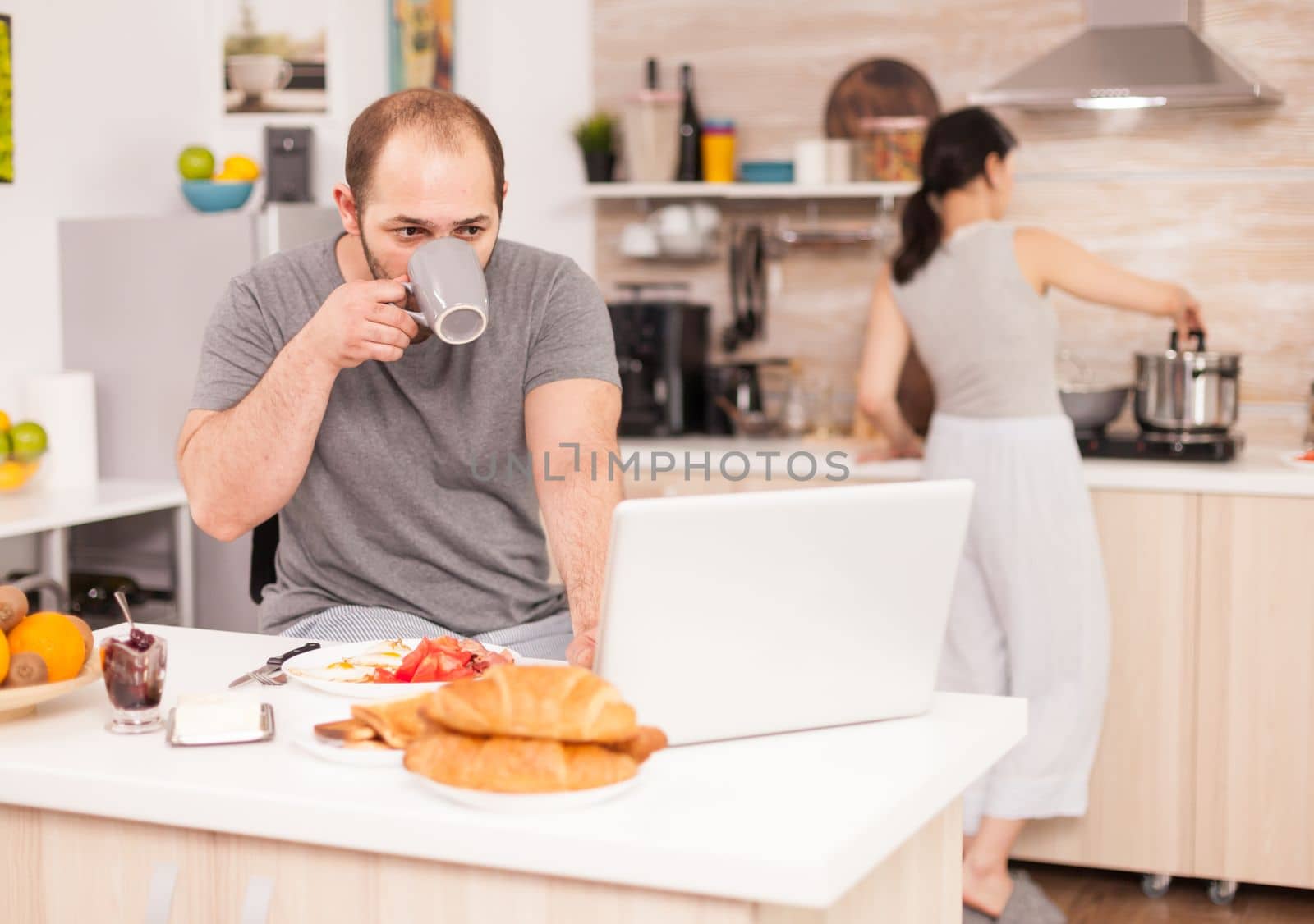 Entrepreneur working on laptop in kitchen and drinking coffee while wife is cooking breakfast. Freelancer work online via internet using modern digital technology