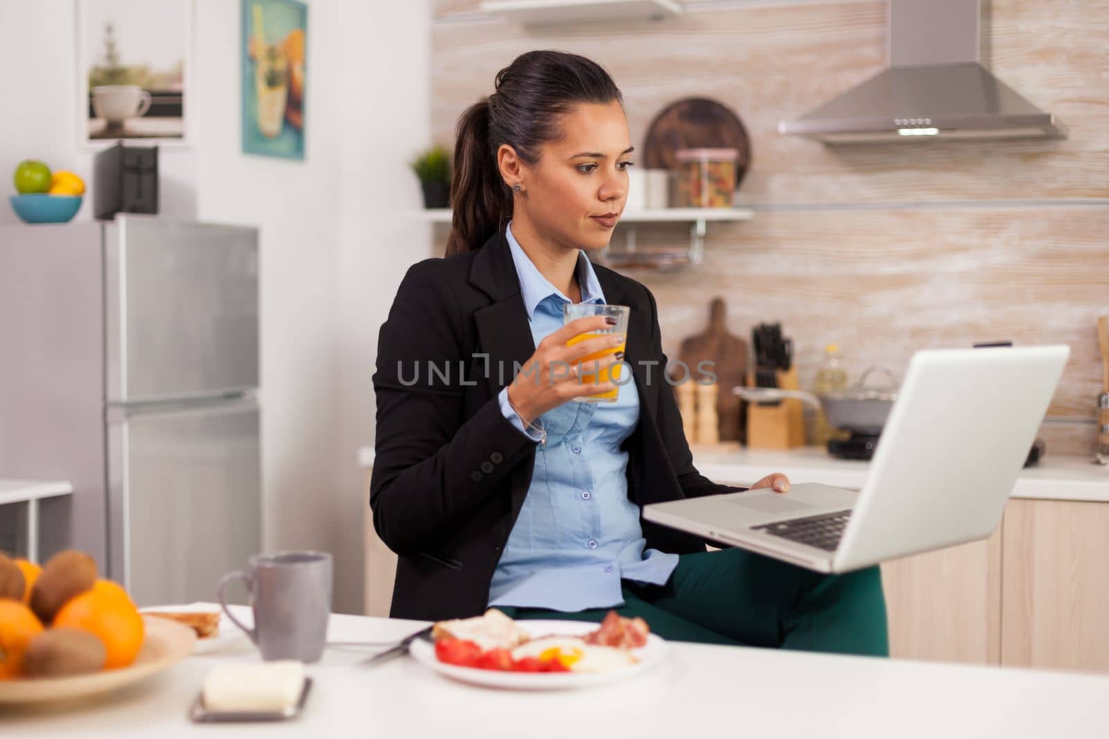 Business woman taking a break and relaxing watching a video clip, drinking orange juice. Young freelancer in the kitchen talking on a video call with her colleagues from the office, using modern internet technology