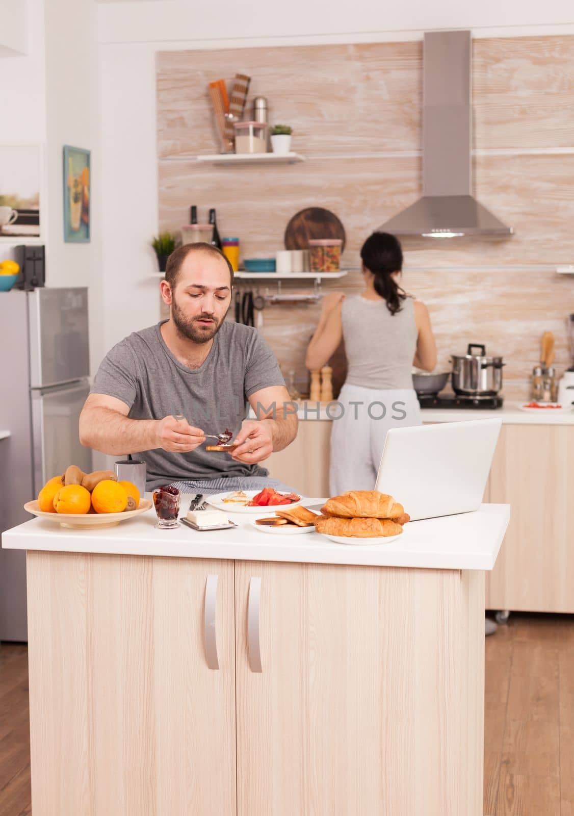 Independence worker eating breakfast and working from home while his wife cooks. Freelancer working remote, talking in videoconference video call online web internet meeting from home, communication device