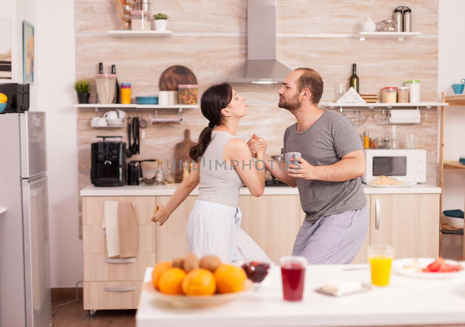 Cheerful couple in pajamas dancing in kitchen listening music during breakfast. Carefree husband and wife laughing, singing, dancing listening musing, living happy and worry free. Positive people. Handheld shot