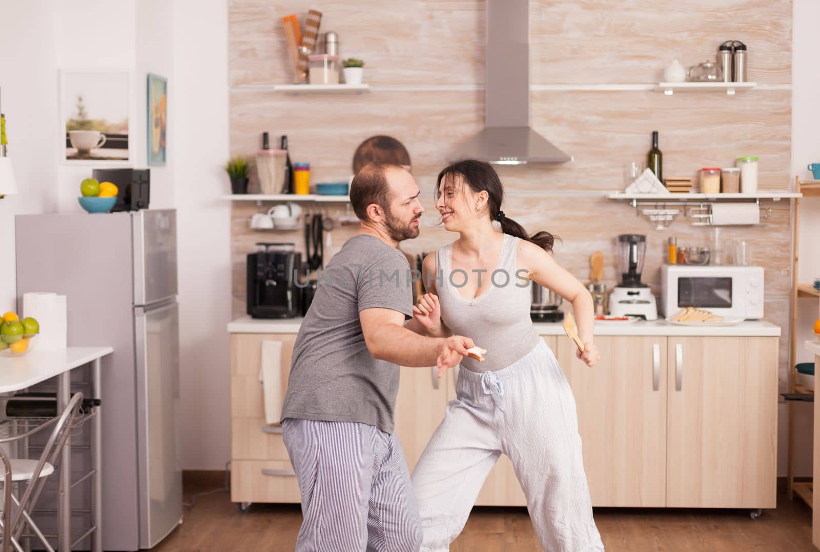 Positive cheerful crazy couple dancing while having breakfast in kitchen wearing pajamas. Carefree wife and husband laughing having fun funny enjoying life authentic married people positive happy relation