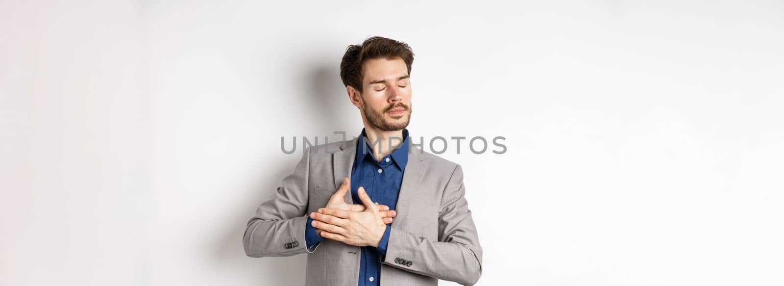 Romantic bearded man in stylish suit holding hands on heart, close eyes and remember something with tender feelings, standing nostalgic on white background.