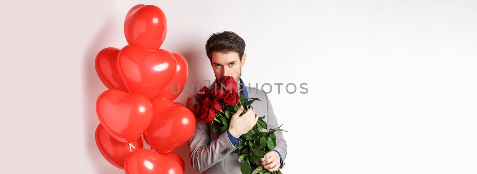 Romantic man smell bouquet of red roses and looking passionate at camera. Boyfriend in suit going on Valentines date with gifts and heart balloons, white background.