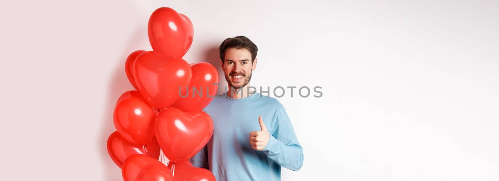 Smiling caucasian man standing with heart balloon, prepare surprise for lover on Valentines day, showing thumbs up and looking at camera, white background.