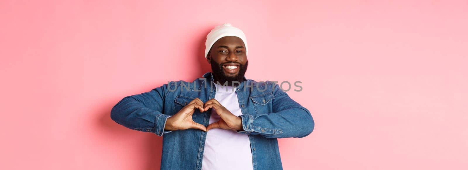 Happy african-american man showing heart sign, I love you gesture, smiling at camera, pink background by Benzoix