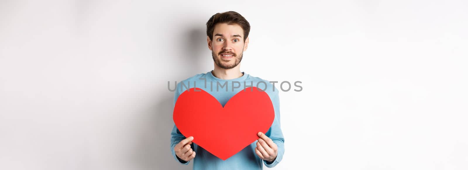 Relationship and love concept. Handsome caucasian man in sweater holding big red valentines day heart cutout and smiling, confessing on date, standing over white background by Benzoix