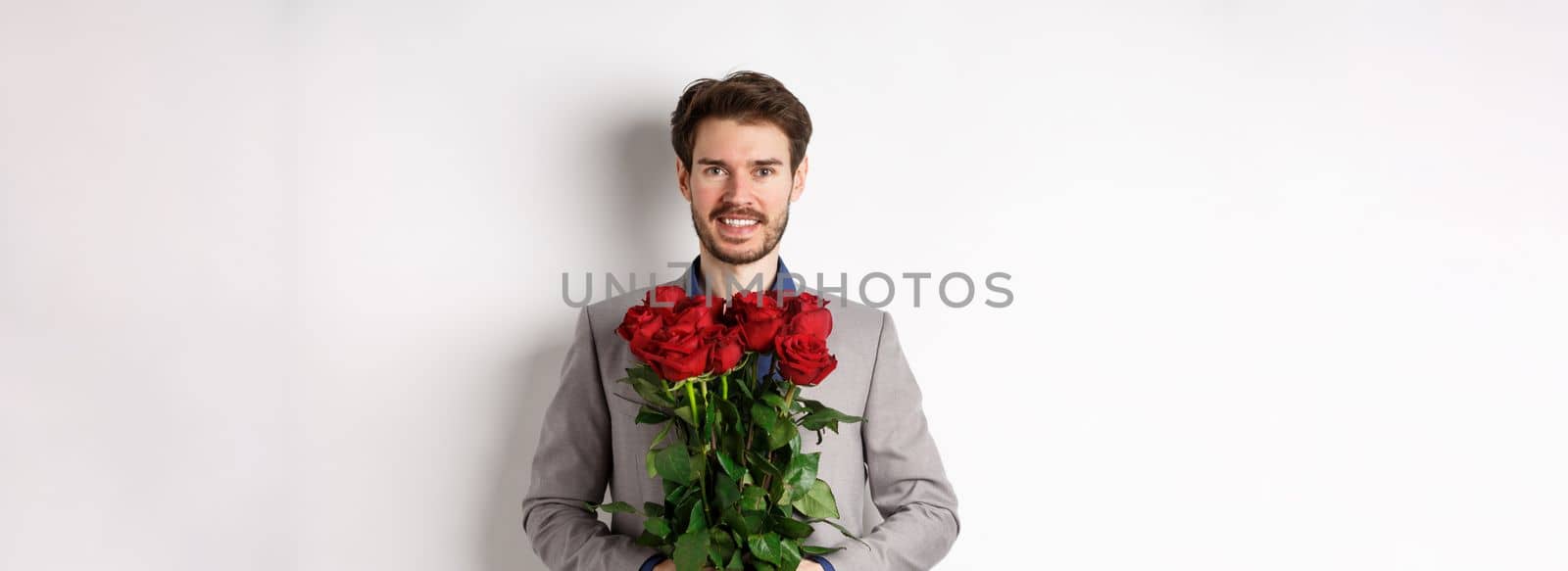 Handsome young man in suit standing with gift on valentines day, holding red roses bouquet and smiling at lover, standing over white background by Benzoix