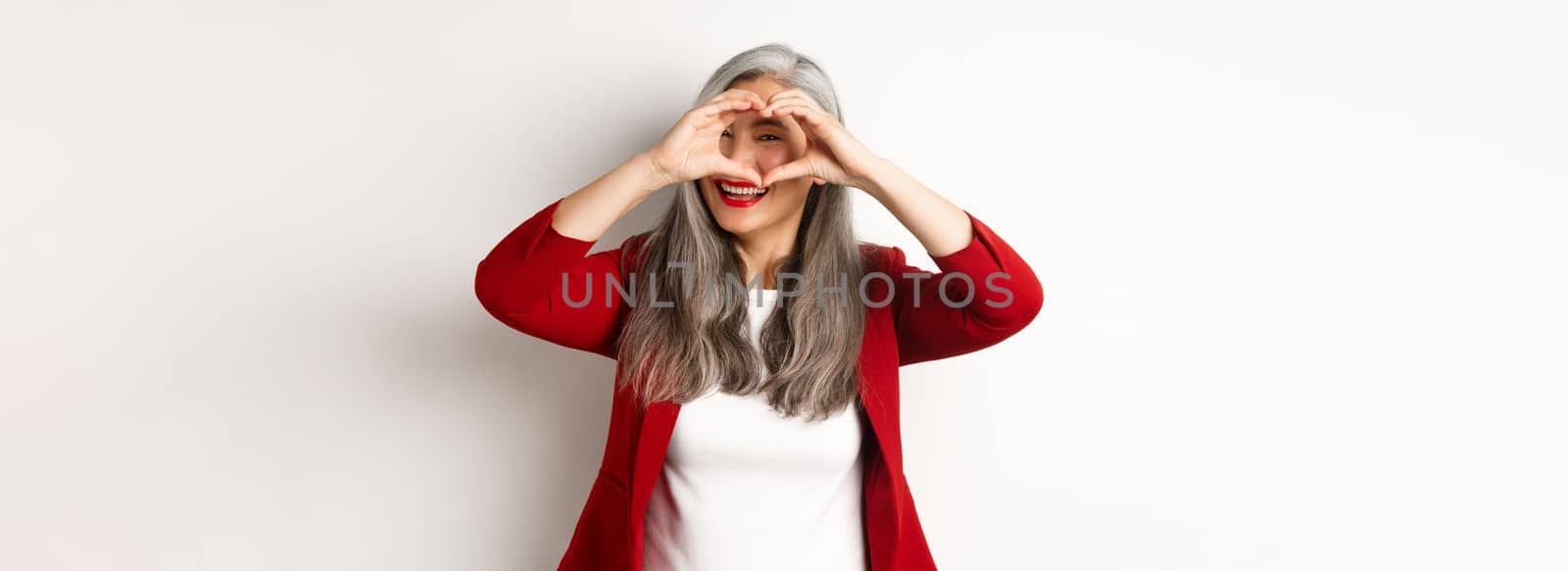 Lovely asian middle-aged woman with grey hair, wearing red blazer, showing heart sign and peeking throught it, I love you gesture, standing over white background by Benzoix