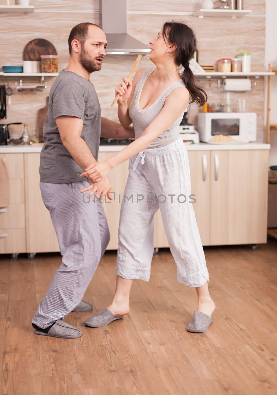 Couple having fun in the kitchen fencing with big spoons during breakfast wearing pajamas. Cheerful carefree joyful funny lovers, fighting wooden spoon, bonding game fight swordplay, happy lifestyle