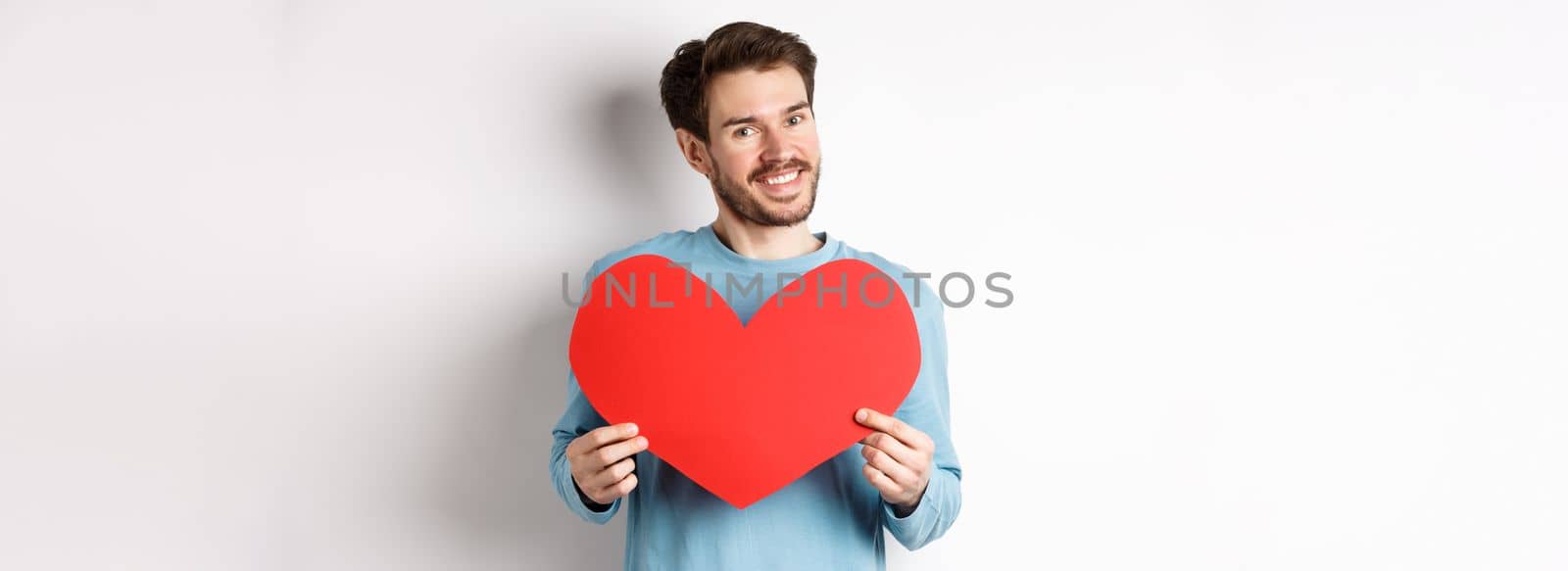 Handsome european man in sweater saying I love you, boyfriend standing with valentines day red heart, posing over white background by Benzoix