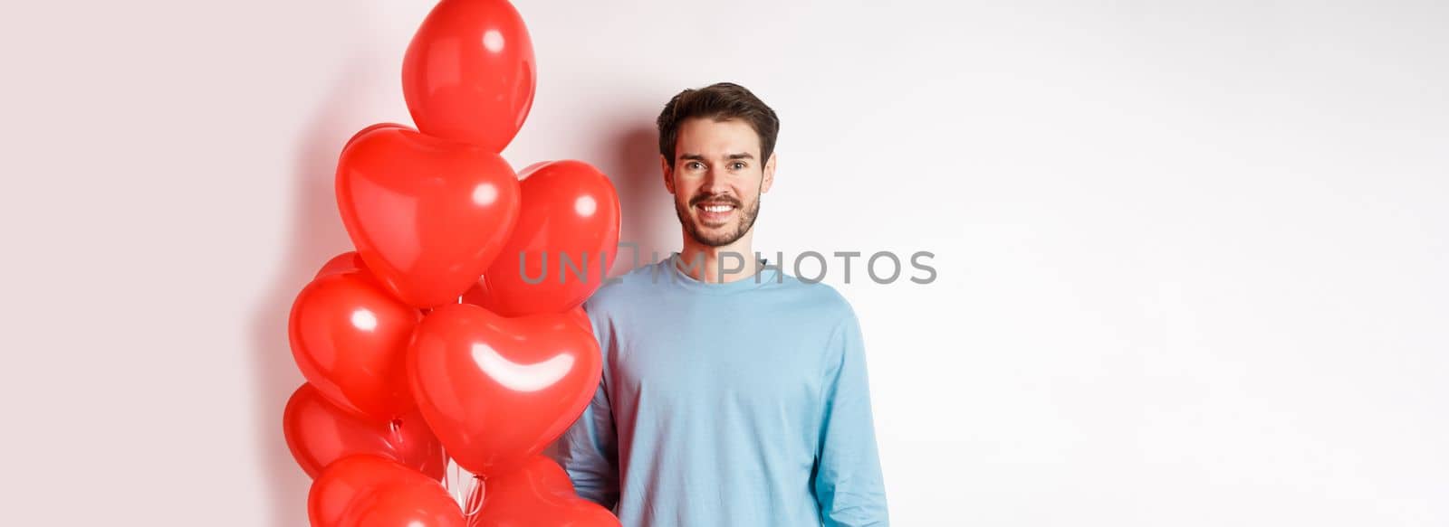 Smiling young man standing with heart balloons and looking happy, celebrating valentines day, bring romantic present to lover, standing over white background.