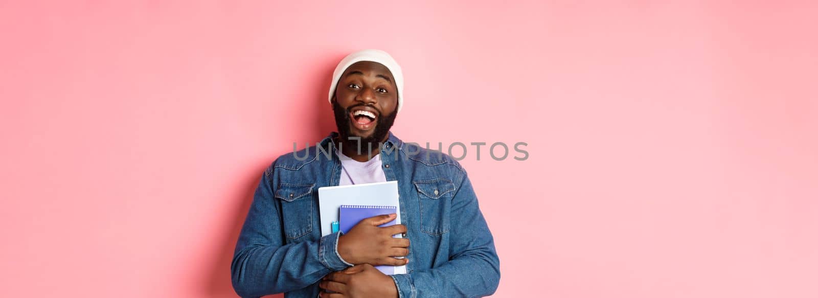 Education. Happy african-american male student in beanie holding notebooks, studying courses, smiling at camera, standing over pink background by Benzoix