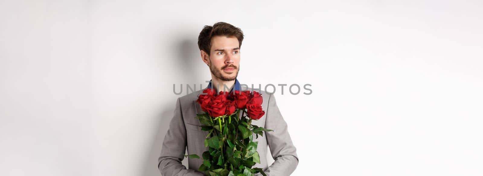 Handsome boyfriend in suit going on romantic date, holding bouquet of red roses and looking left thoughtful, standing over white background.