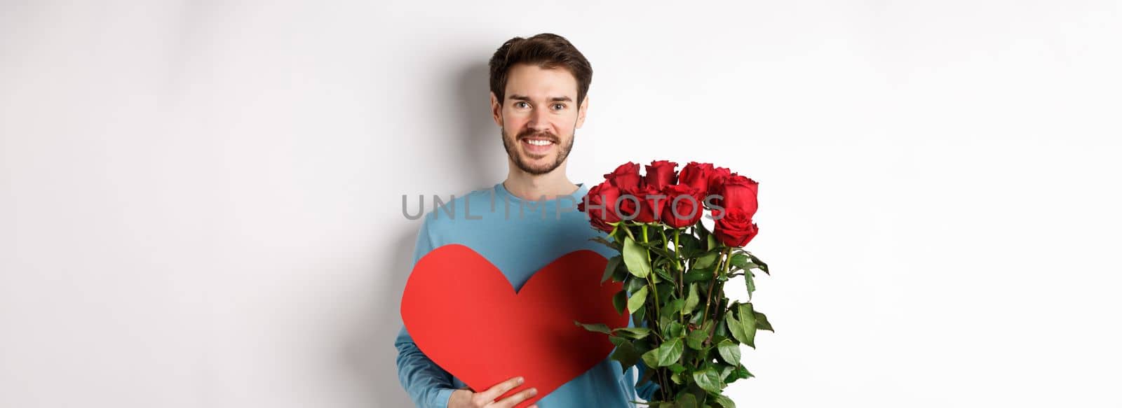 Romantic people. Handsome smiling man holding bouquet of roses and big red heart, going on Valentines day date with girlfriend, standing over white background by Benzoix