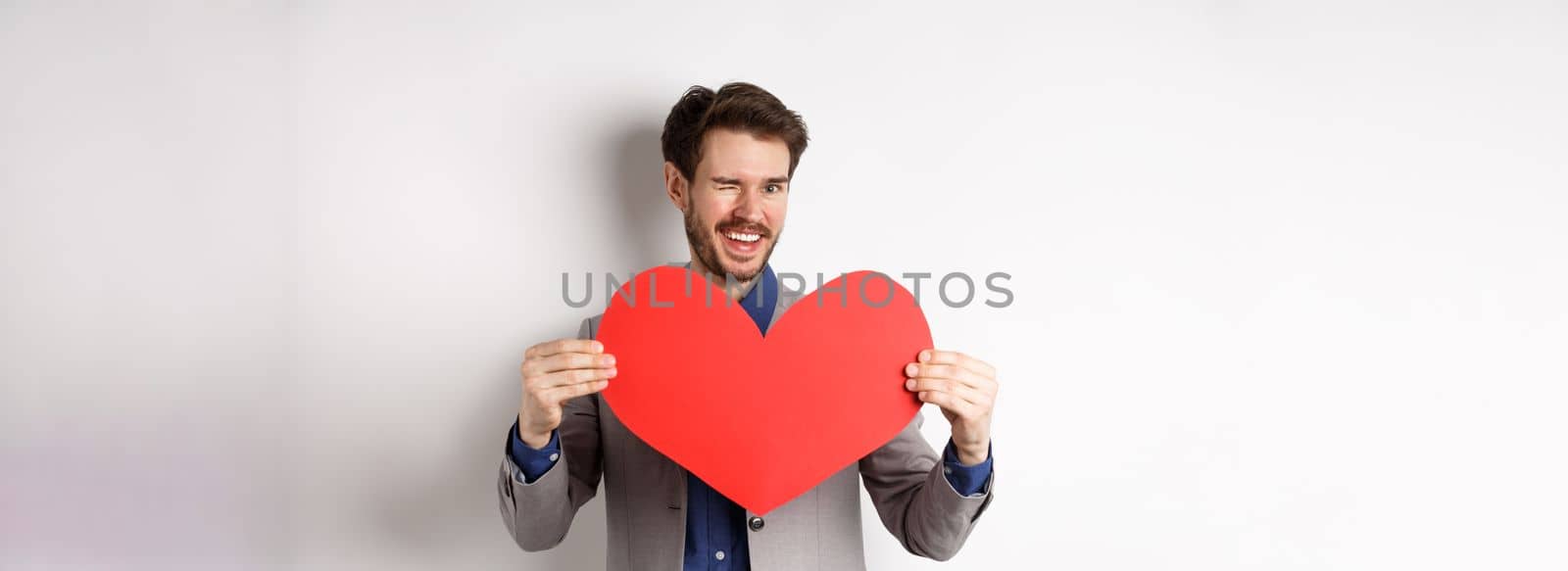 Charismatic young man winking and smiling, showing big red heart cutout for Valentines day date, say love you to lover, standing over white background.