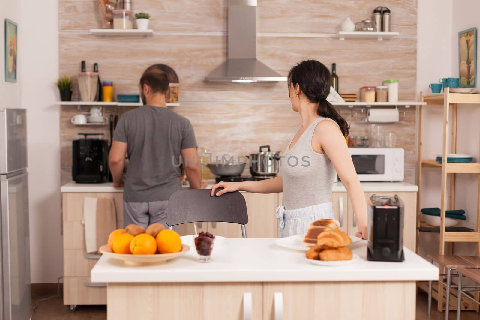 Cheerful wife talking with her husband in the kitchen while toasts the bread for breakfast. Young couple in the morning preparing meal together with affection and love
