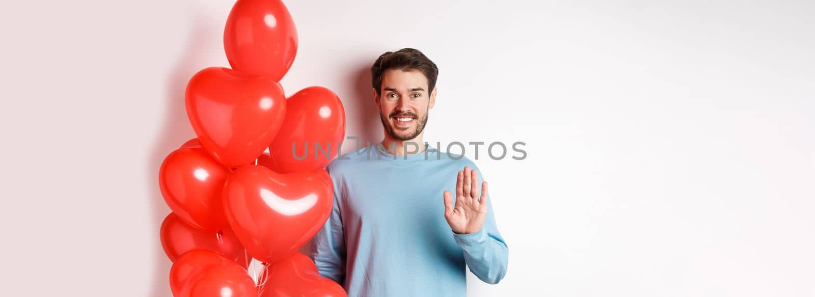 Handsome boyfriend saying hello, bring romantic red heart balloons on date, waving hand and smiling, standing over white background by Benzoix