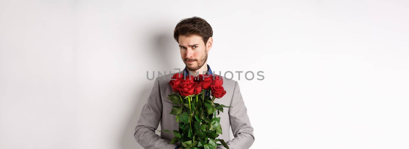 Indecisive bearded man in suit, holding flowers roses and looking at camera unsure, going on romantic date, standing over white background by Benzoix