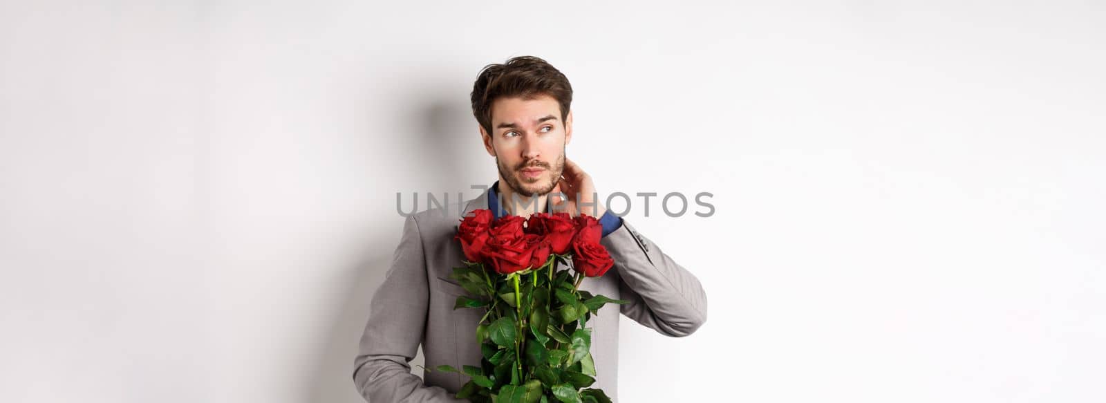 Pensive young man in suit holding bouquet of flowers, waiting for date on Valentines day, standing over white background by Benzoix