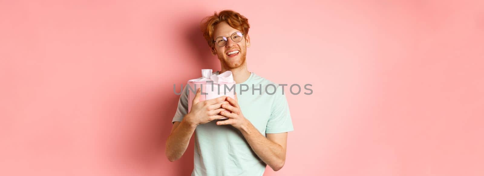 Valentines day and romance concept. Happy redhead boyfriend receive romantic gift, hugging box with present and saying thank you, smiling grateful, pink background by Benzoix