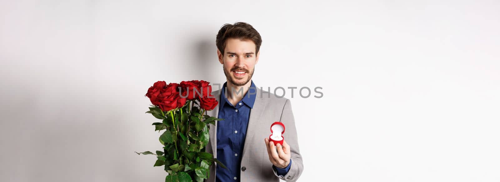 Smiling boyfriend making a marriage proposal, standing with engagement ring and bouquet of red roses, going on romantic Valentines day date, white background by Benzoix