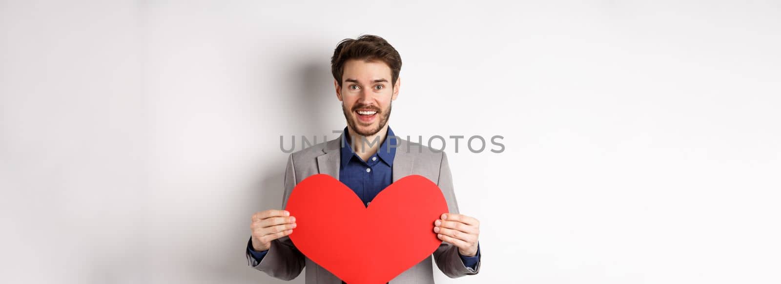 Happy man confessing in love, showing heart cutout and smiling at camera, standing in suit on romantic date with lover, white background by Benzoix