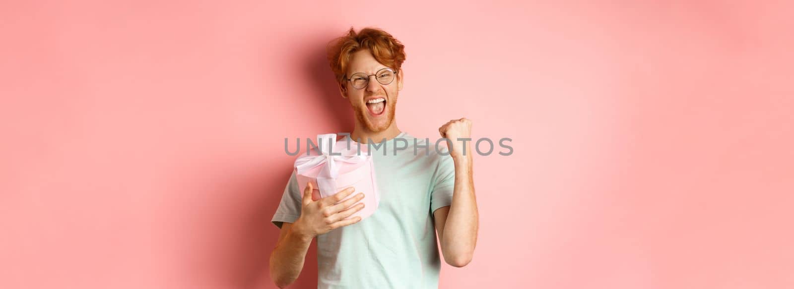 Valentines day and romance concept. Cheerful redhead guy making fist pump and scream yes with joy, holding gift box, smiling, standing over pink background.