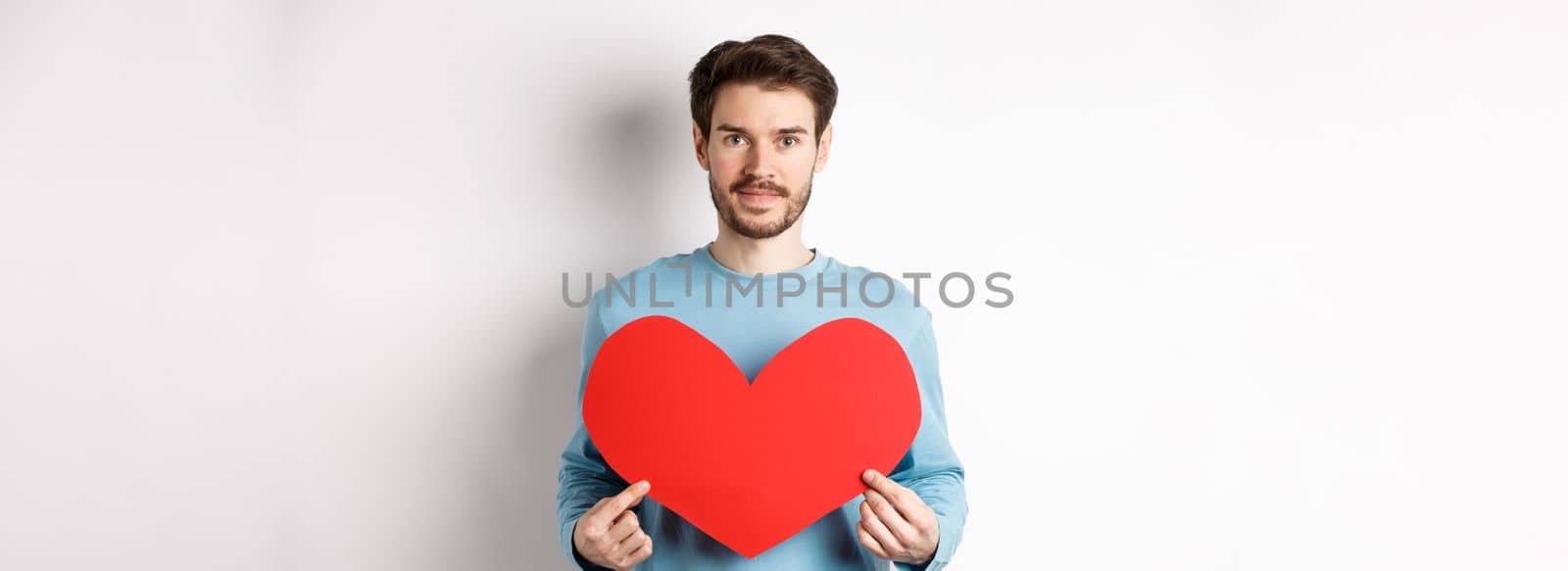 Smiling young man holding valentines heart cutout and looking at camera, waiting for true love girlfriend, standing over white background.