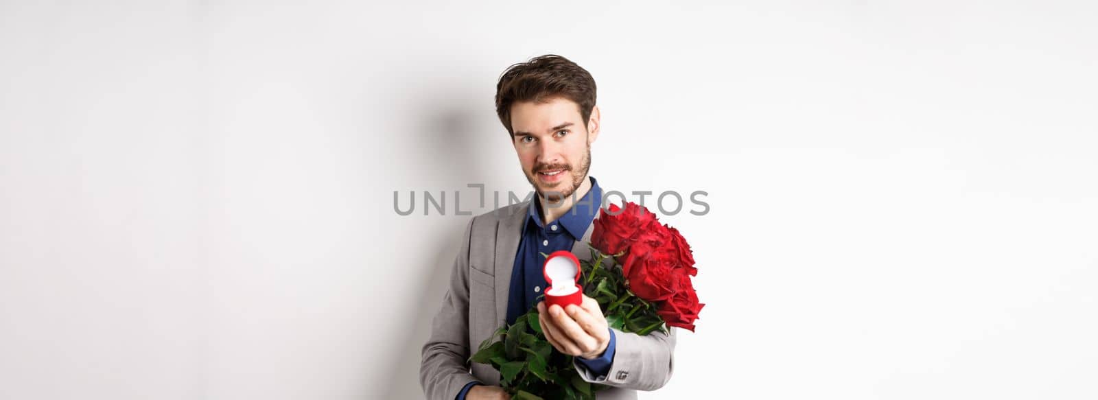 Romantic man with boquet of red roses asking to marry him, holding engagement ring and looking confident at camera, standing in suit over white background by Benzoix