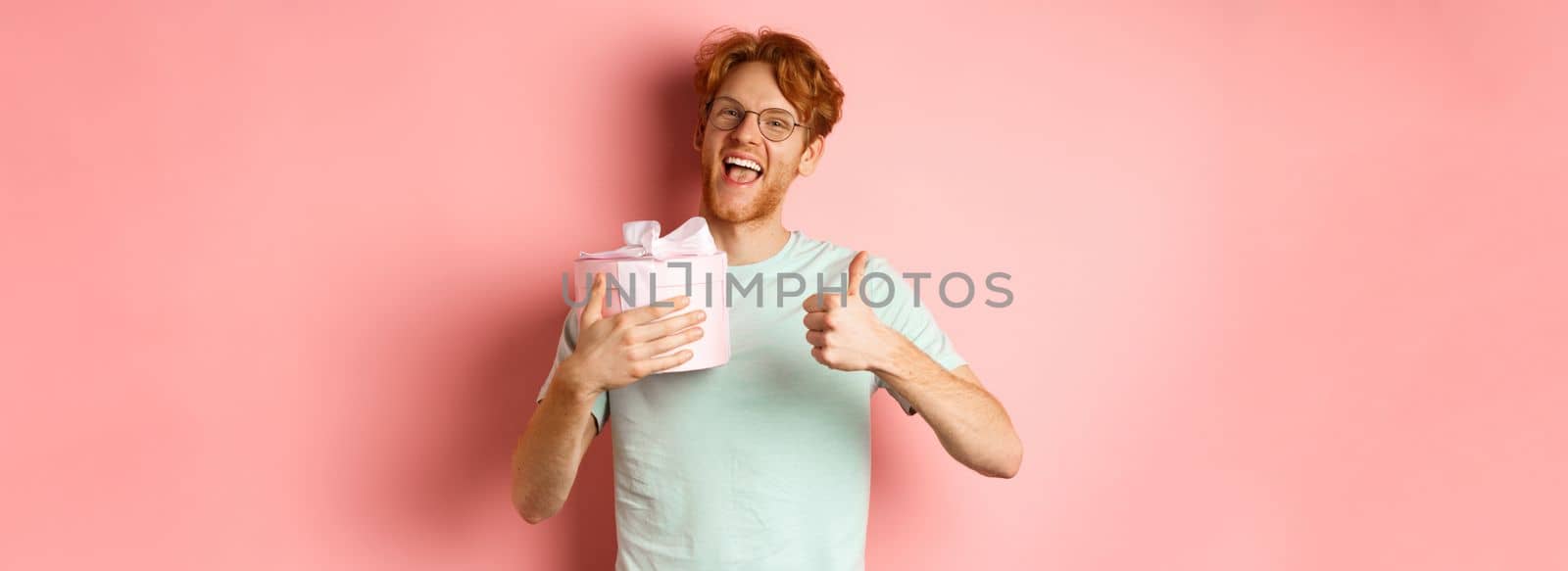 Valentines day and romance concept. Cheerful young man holding box with gift and showing thumbs-up, thanking for present, standing over pink background.