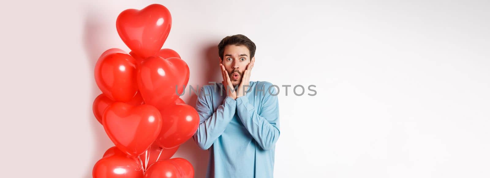 Valentines day. Image of young man standing near hearts balloons with shocked face, staring startled at camera, white background.