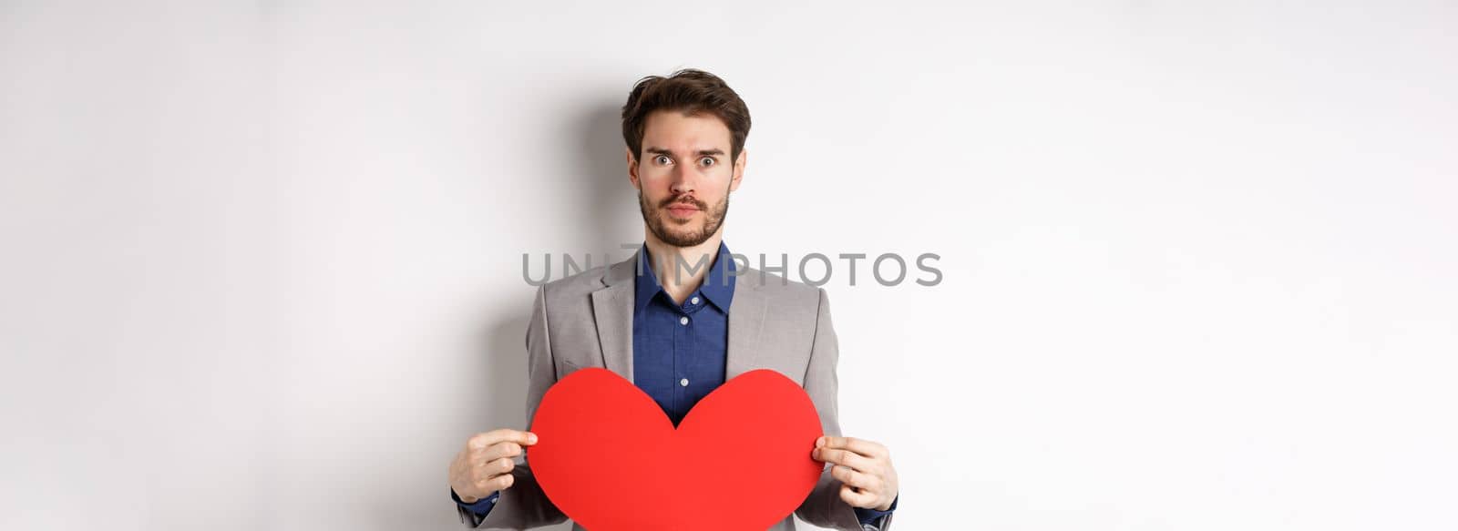 Excited caucasian man in suit looking at camera, holding big red heart cutout on valentines day, standing over white background by Benzoix