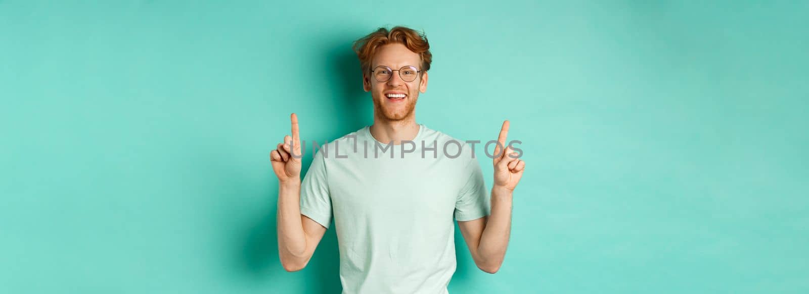 Image of handsome bearded man with red hair, wearing t-shirt and glasses, smiling happy and pointing fingers up, showing promo offer, standing over turquoise background by Benzoix