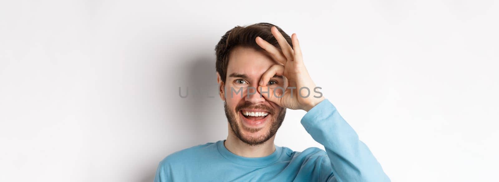 Excited smiling man looking through okay sign with happy face, approve and like promo offer, standing over white background.