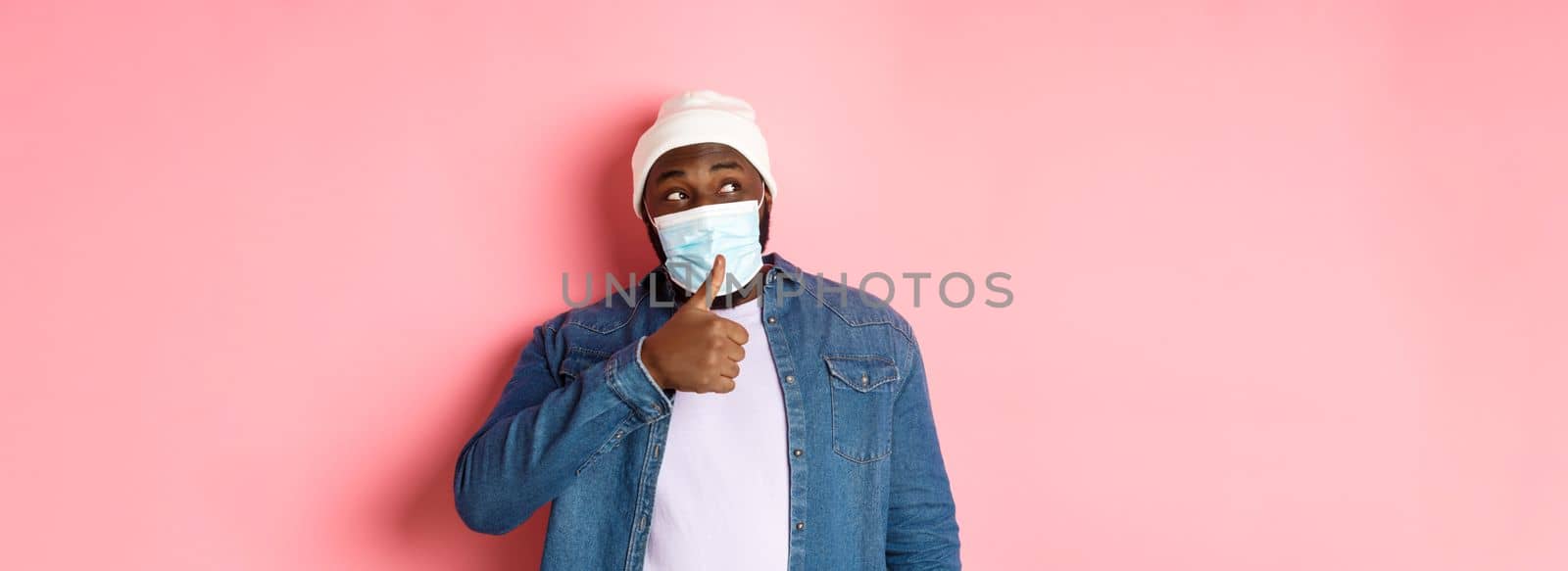 Covid-19, lifestyle and quarantine concept. Cheerful Black man showing thumbs-up and looking left, praising promo offer, approve and like, standing over pink background by Benzoix