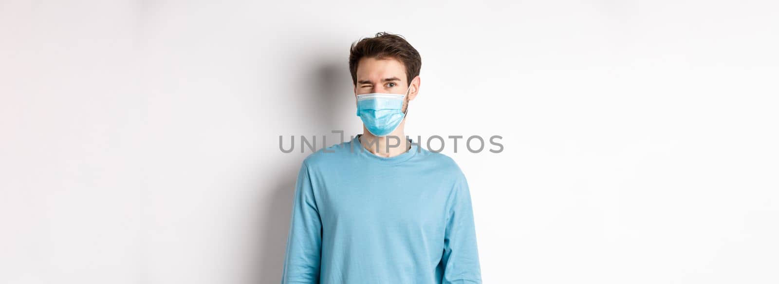 Covid-19, pandemic and social distancing concept. Cheerful caucasian man in face mask winking at camera, looking happy, standing over white background.