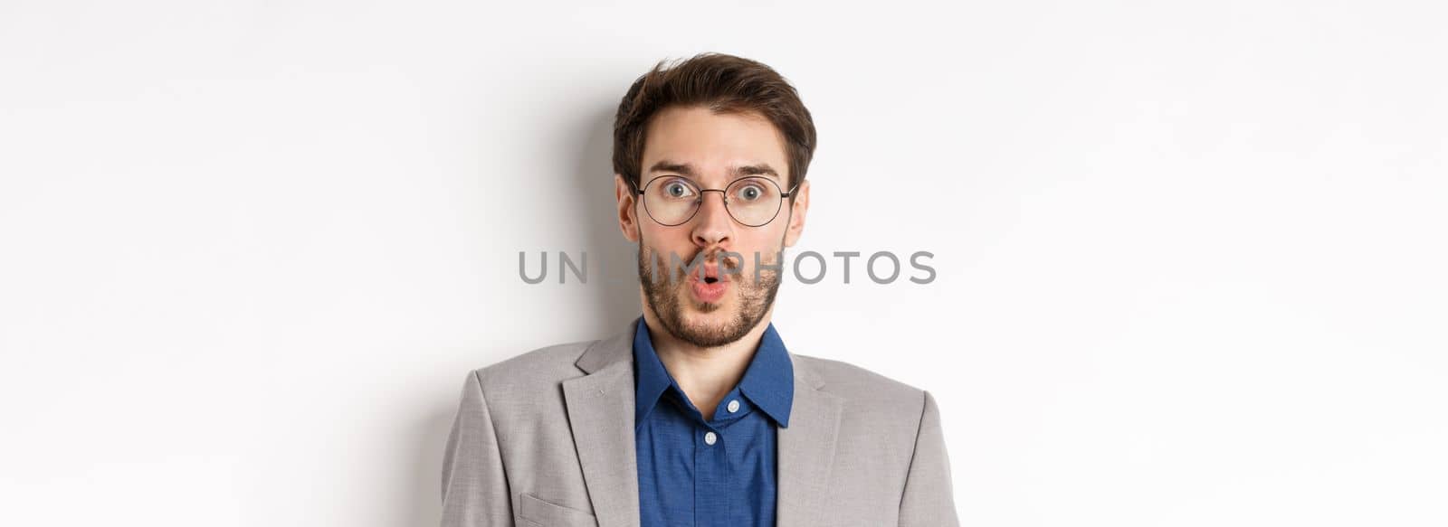 Close up portrait of excited office worker in glasses and suit saying wow, staring amazed at camera, standing against white background.