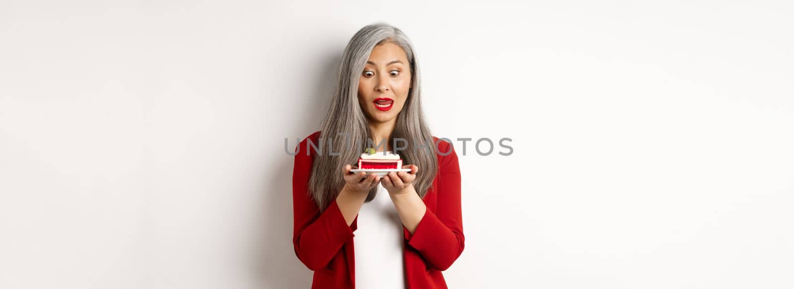 Image of funny asian senior woman looking tempted at piece of cake, desire to bite dessert, standing over white background.
